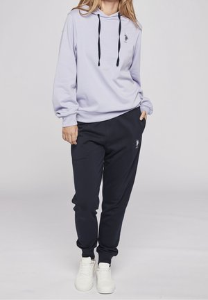 Frau trägt hellvioletten Hoodie mit schwarzen Kordeln und schwarze Jogginghose, steht mit einer Hand in der Tasche vor einfarbigem Hintergrund.