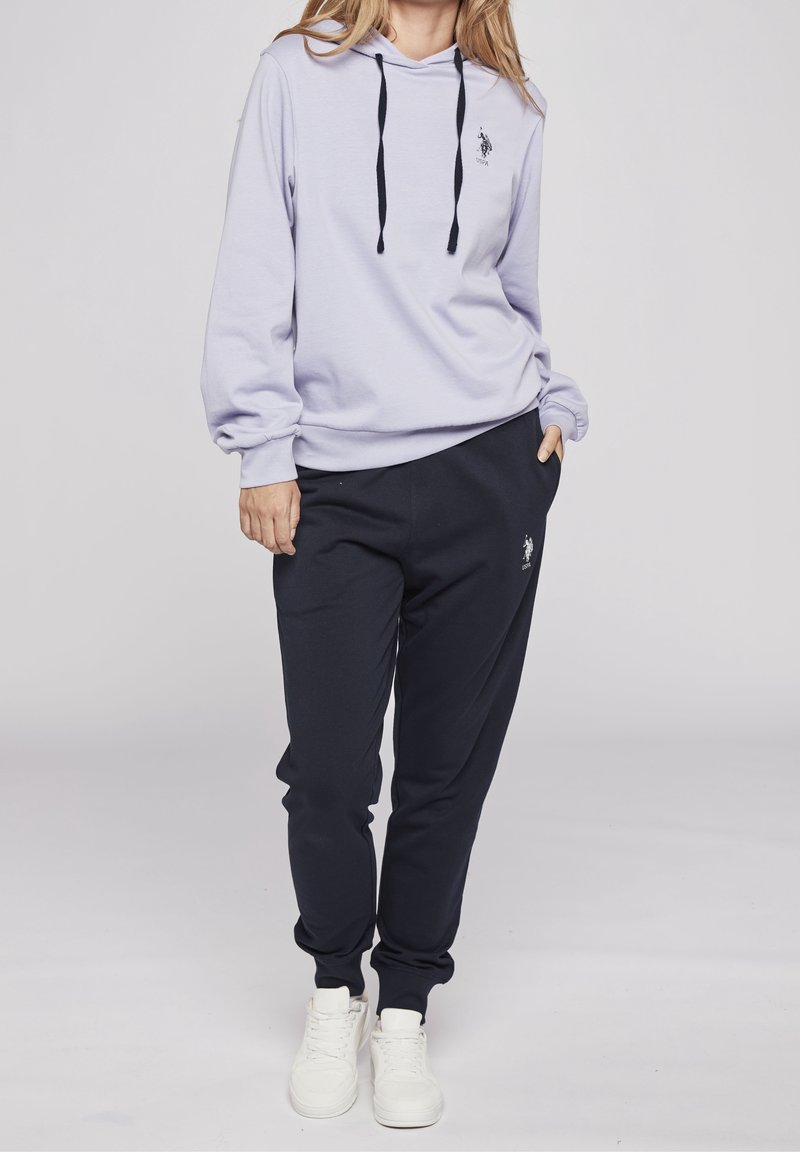 Femme portant un hoodie violet clair avec cordons noirs et un pantalon de jogging noir, debout avec une main dans la poche sur un fond uni.