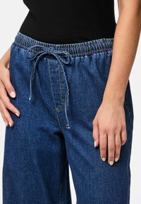 Tumši zili džinsu bikses ar elastīgu jostu un izvilkšanas auklu. Iespējām divas sānu kabatas un brīvu, plašu kāju piegriezumu.