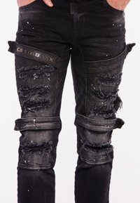 Schwarze Jeans mit used-Effekten, die Risse und fransige Kanten aufweisen. Ein strukturierter Logo-Patch ist am Oberschenkel angebracht, ebenso wie Farbkleckse.