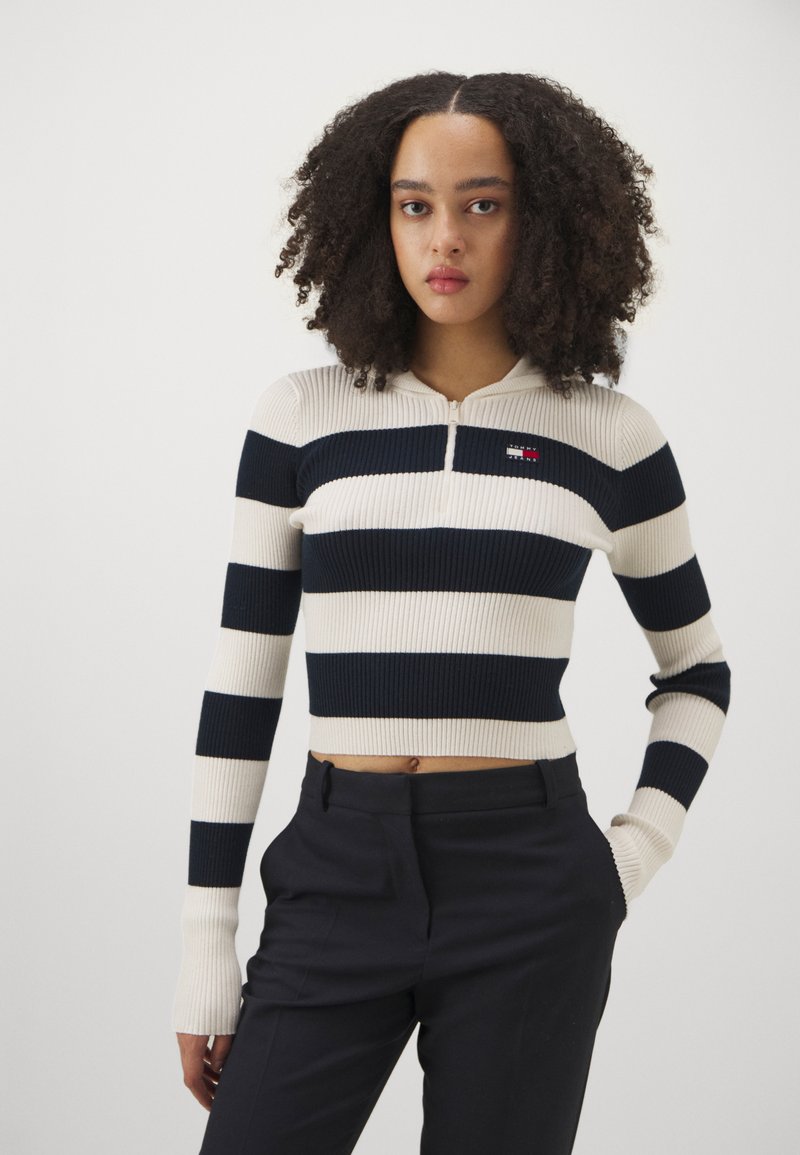 Varrukatega lühikeste õlgadega kampsun, millel on tumesinised ja kreemjad horisontaalsed triibud, esine lukuga ning rinnal väike logo silt.