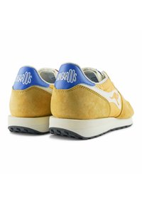 Scarpe sportive con suede giallo, dettagli bianchi e un logo blu sul tallone. Presentano una robusta intersuola di colore crema e una suola nera.