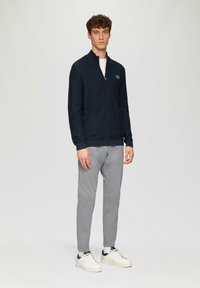 Marineblauwe zip-up trui met hoge kraag, geribbelde manchetten, gedragen over een wit shirt. Gecombineerd met lichtgrijze broeken en witte sneakers.