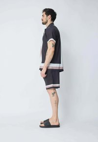 Chemise noire à manches courtes avec des accents blancs et bordeaux, associée à un short assorti. Portée avec des sandales noires à enfiler, montrant des tatouages visibles sur les jambes.