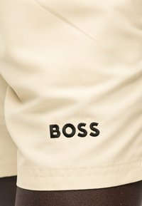 Μπεζ ύφασμα με τη λέξη «BOSS» κεντημένη με μαύρη κλωστή κοντά στην άκρη του υλικού.