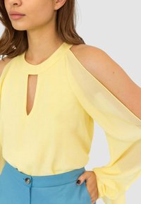 Blusa gialla con scollo a goccia e design a spalle scoperte, caratterizzata da maniche trasparenti e una texture liscia. Abbinata a pantaloni blu.