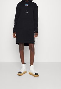 Svart huvtröja med framficka, prydd med en liten logotag. Bärs med vita höga sneakers med svarta accenter och gummisula.