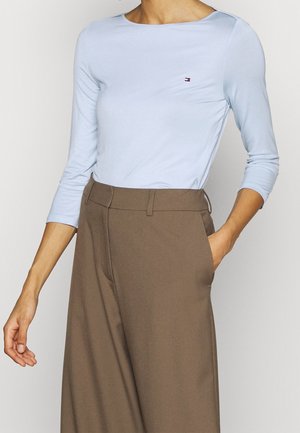 Frau trägt ein hellblaues Langarmshirt mit kleinem Logo und hoch taillierte, lockere braune Hose, eine Hand in der Tasche.