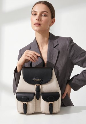 Frau im grauen Blazer hält einen beigen und schwarzen DIANA&CO Firenze Rucksack mit Schnallenverschlüssen auf weißem Hintergrund und Oberfläche.