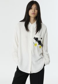 Chemise oversized blanche à texture avec un col à boutonnage; présente un patch brodé noir et jaune sur la poitrine gauche.