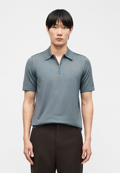 Άνδρας που φοράει γκρι πόλο μπλουζάκι με κοντά μανίκια, φερμουάρ μπροστά και σκούρο καφέ παντελόνι, στεκόμενος μπροστά σε απλό λευκό φόντο.