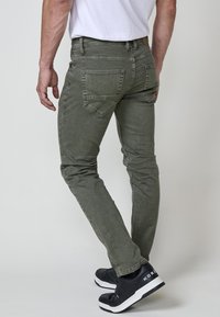Olivno zeleni slim-fit džins s petimi žepi, vidnim šivom in subtilnimi detajli obrabe. V kombinaciji z črnimi športnimi copati s belimi detajli.