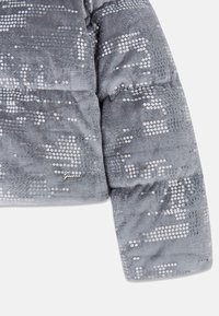 Graue Steppjacke mit einer strukturierten, samtigen Oberfläche, die silberne Pailletten in einem gemusterten Arrangement aufweist. Enthält ein "Guess"-Logo-Detail.