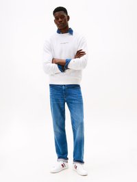 Felpa bianca con logo, indossata sopra una camicia di jeans, abbinata a jeans blu e sneakers bianche con strisce rosse e blu navy.
