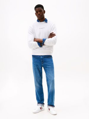 Sweatshirt branco com logotipo, sobreposto a uma camisa de ganga, combinado com jeans azuis e ténis brancos com riscas vermelhas e azuis-marinho.