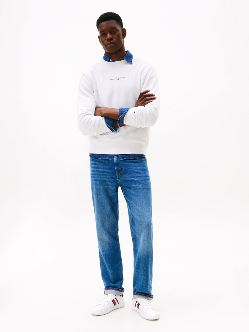 Felpa bianca con logo, indossata sopra una camicia di jeans, abbinata a jeans blu e sneakers bianche con strisce rosse e blu navy.