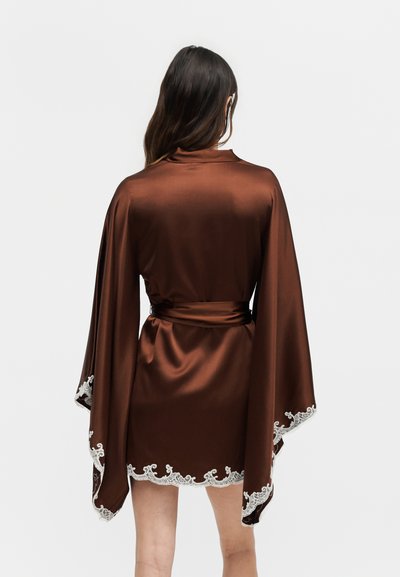 Γυναίκα που φορά καφέ σατέν ρόμπα με φαρδιά μανίκια και λευκή δαντέλα στα μανσέτα και το τελείωμα, στέκεται με την πλάτη προς ένα απλό φόντο.