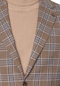 Brązowa marynarka w kratę z beżowym golfem. Wyróżniające się detale to niebieskie i białe akcenty w wzorze.