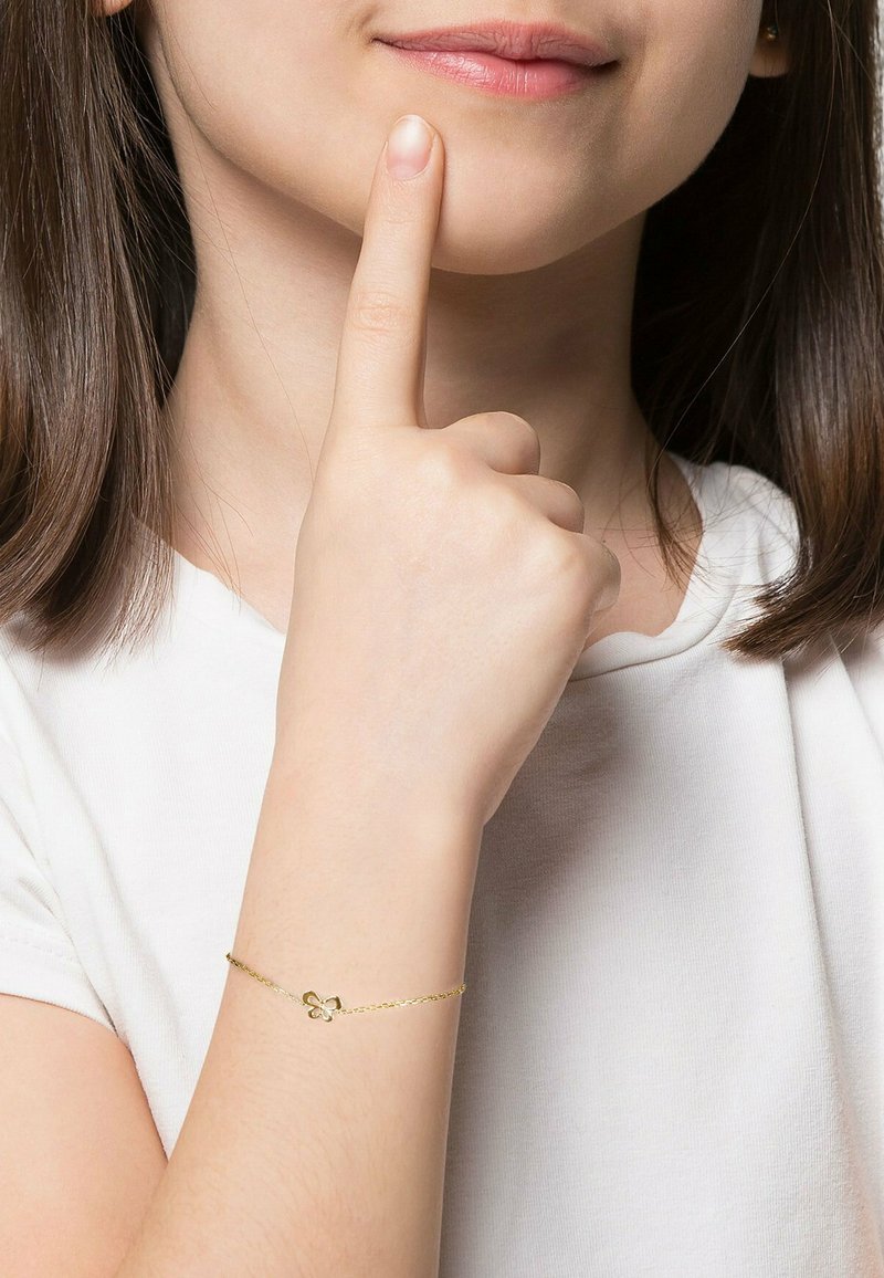 Junge Person trägt ein weißes Hemd mit einem goldenen Schmetterlingsarmband und berührt mit dem Zeigefinger das Kinn in einer nachdenklichen Geste.