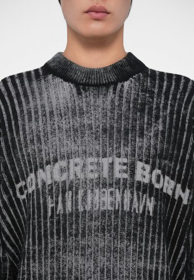 Melns un pelēks ribots džemperis ar apaļu kakla izgriezumu, kurā uzraksts "CONCRETE BORN" ir vieglākā krāsā, mīksta tekstūra.