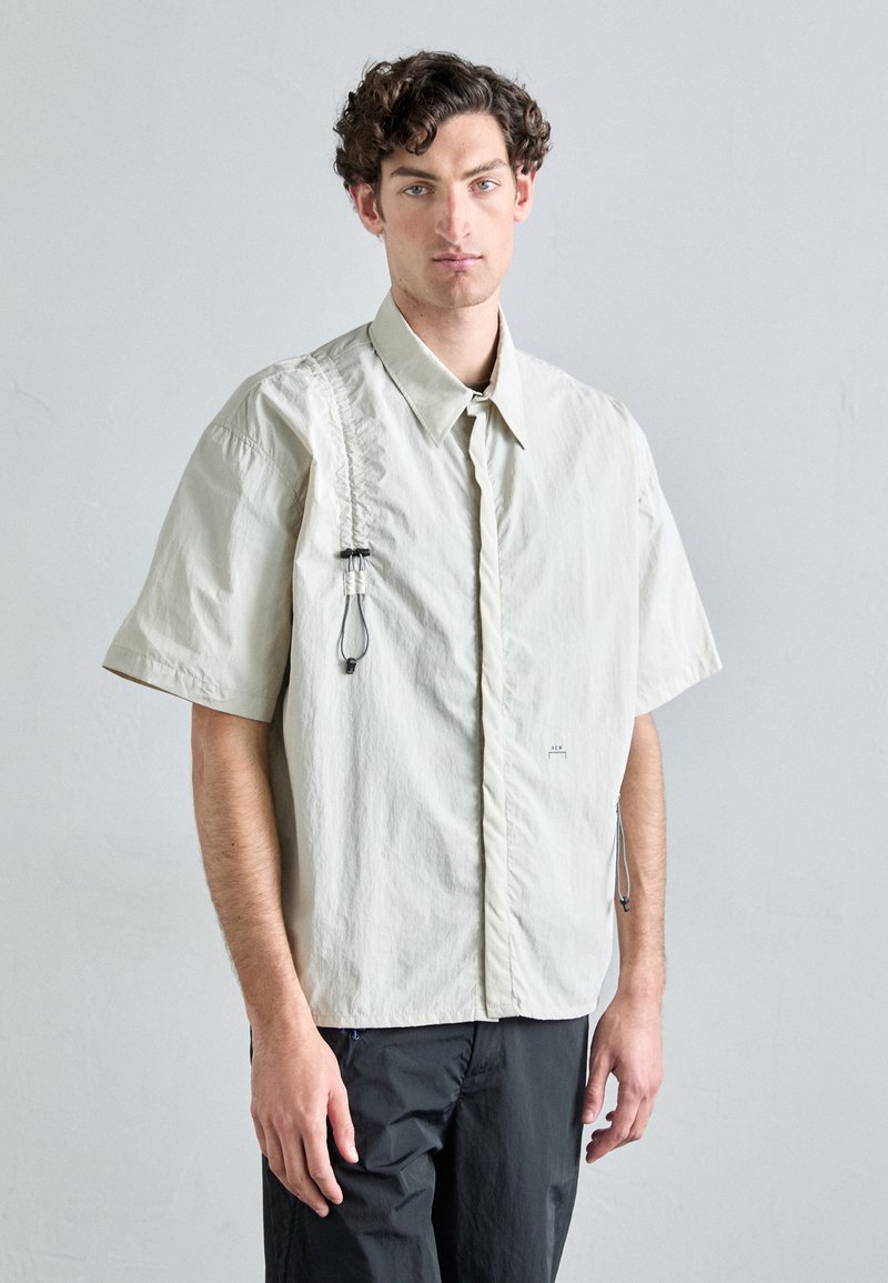 Chemise beige clair à manches courtes avec une fermeture à boutons, en tissu texturé, détail de boucle décorative sur le côté et petit logo près de l'ourlet.