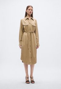 Μπεζ πουκαμισοφόρεμα με δύο τσέπες στο στήθος, ζώνη στη μέση, μακριά μανίκια, κουμπωτό μπροστινό μέρος και στρογγυλεμένη γραμμή στο τελείωμα. Μαύρα σανδάλια με λουράκια.