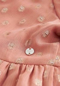 Gros plan sur un tissu rose présentant un motif texturé de petits carrés dorés, avec un bouton argenté inscrit "M+V" attaché par un fil.