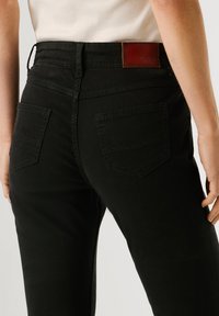 Gros plan sur une personne portant un jean noir taille haute avec poches arrière et écusson en cuir marron de la marque, assorti à un haut blanc.
