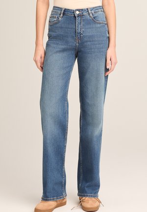 Frau trägt hoch taillierte, gerade geschnittene blaue Jeans mit Fronttaschen und hellbraunen Wildlederschuhen mit Schnürsenkeln.