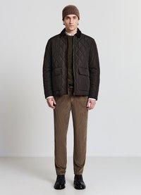 Steppjacke in Dunkelbraun mit Kragen, zwei Vordertaschen, darüber ein strukturiertes grünes Sweatshirt, kombiniert mit brauner Cordhose und schwarzen Stiefeln.