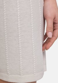 Pull en tricot de couleur beige clair, présentant des rayures verticales texturées et un ourlet côtelé. Posé à la main sur le côté, montrant des détails de couture.