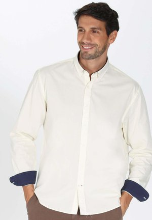 Chemise beige clair à manches longues avec un col boutonné. Les poignets présentent un accent bleu marine contrastant. Texture lisse, design minimal.
