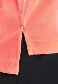 Top deportivo coral con mangas cortas, dobladillo cosido y detalle de abertura lateral. El material parece suave y elástico, combinado con tela negra en la parte inferior.