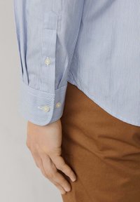 Camisa de rayas azul claro con botones blancos y detalle en los puños, combinada con pantalones de tela marrón. Textura suave y corte entallado.
