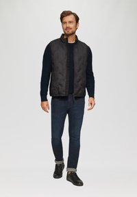 Gilet matelassé noir sur un pull en maille bleu marine, associé à un jean foncé et des baskets noires. Le gilet présente une fermeture éclair à l'avant et un col haut.