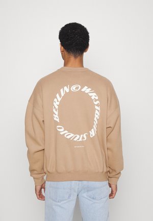 Personne aux cheveux courts et bouclés portant un sweat-shirt beige avec un texte blanc circulaire dans le dos et un jean bleu clair, vue de dos.