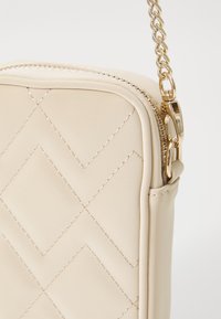 Prošívaná béžová crossbody taška s hladkou texturou z umělé kůže, zlatým řetízkovým popruhem a zipovým uzávěrem s kovovými detaily.