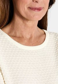 Cremefarbener Strickpullover mit strukturierten Oberflächen und einem breiten, abgerundeten Ausschnitt, der eine Nahaufnahme des Kragens und des Nackenbereichs zeigt.