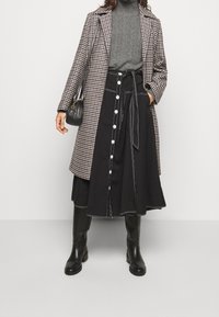Manteau à carreaux marron et blanc porté sur un col roulé gris, associé à une jupe noire ornée de boutons et d'une ceinture à nouer, le tout complété par des bottes noires.