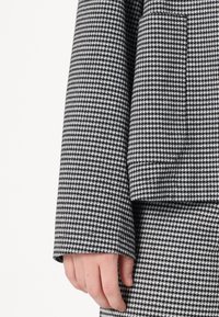 Svart og hvitt houndstooth-mønstrete stoff med glatt tekstur, med vid erm og lomme detaljer.