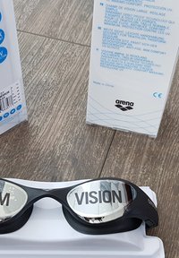 Zwarte zwembril met spiegelende lenzen met de tekst "VISION," met een rubberen frame voor comfort, rustend op een witte doos.