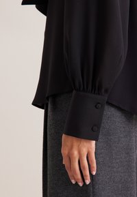 Schwarze Bluse mit langen, leicht gepufften Ärmeln. Die Manschetten haben zwei Knopfakzente. Glattes, leichtes Material; getragen über grauen, strukturierten Hosen.