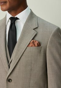 Chaqueta de traje de cuadros gris claro con solapas de muesca, que incluye un pañuelo de bolsillo marrón. Tejido texturizado, complementado con una corbata negra y una camisa blanca.