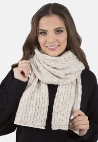 Écharpe en tricot de couleur beige clair, présentant un motif texturé et des bords frangés. Portée lâchement autour du cou du mannequin pour plus de chaleur.
