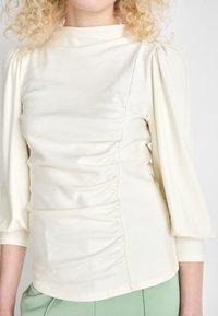 Light cream top with puffed sleeves, featuring horizontal ruching on the front and a high neckline. Smooth, soft fabric with a fitted silhouette.