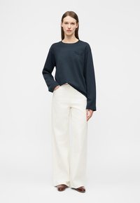 Γυναίκα που φοράει μαύρο πουκάμισο με μακριά μανίκια, λευκό παντελόνι ψηλόμεσο με φαρδιά μπατζάκια και καφέ παπούτσια, στέκεται με το ένα χέρι στην τσέπη.