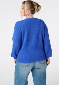 Blauwe gebreide trui met een losse pasvorm, geribde zoom en brede mouwen. Draagt lichtblauwe spijkerbroek en toont een gestructureerde wollen uitstraling.