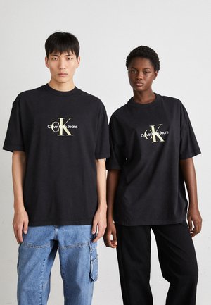 Camisetas de algodón negras de corte oversized, con el logo dorado de "Calvin Klein Jeans", combinadas con pantalones de mezclilla azul y pantalones negros.