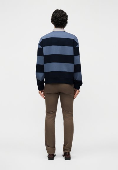 Homme debout, de dos, portant un pull rayé horizontal bleu et noir, un pantalon marron et des chaussures marron foncé.