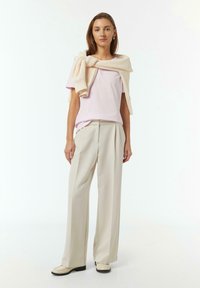 Chemise rose clair à manches courtes, pull en tricot crème drapé sur les épaules, pantalon beige taille haute, mocassins blancs, design minimaliste.